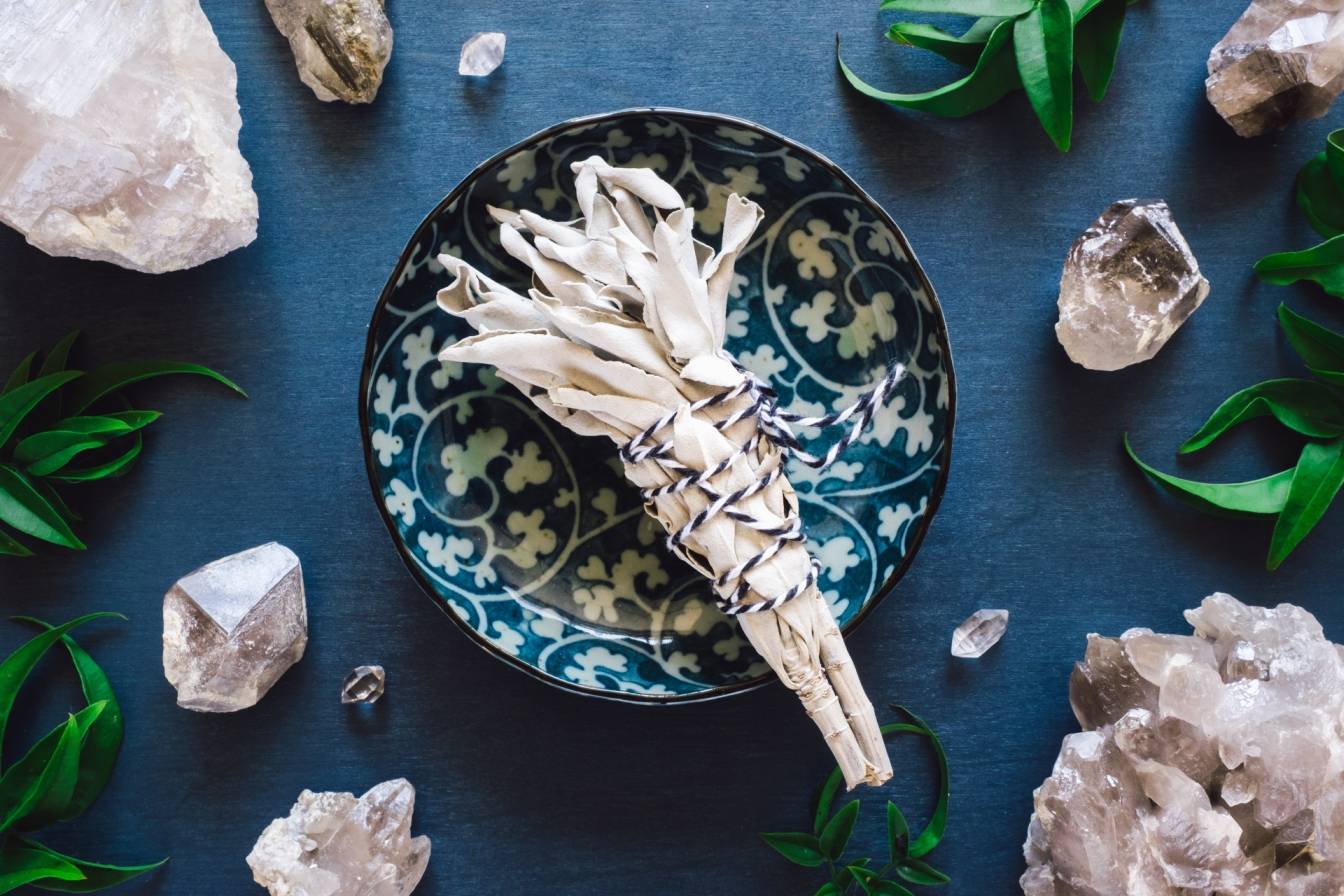 Szałwia jako kadzidło do okadzania i odkażania pomieszczeń leży na talerzu w folklorystyczne wzory na granatowym blacie, wokół leżą liście i minerały.