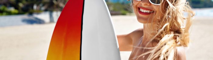 10 rzeczy, które musisz zrobić, aby nie zwariować po urlopie. Uśmiechnięta młoda kobieta w kostiumie kąpielowym i okularach przeciwsłonecznych stoi na plaży z kolorową deską surfingową w ręku.