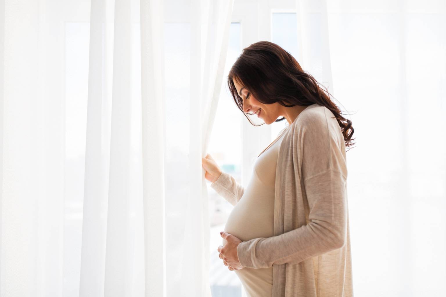 Dlaczego poród boli? Jak rodzić świadomie? Jak pokonać lęk przed bólem podczas porodu? Kobieta w zaawansowanej ciąży stoi przy oknie.