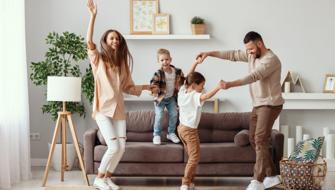 Jak pomóc dziecku rozładować emocje? Felieton Beaty Sadowskiej. Szczęśliwa rodzina - mama, tata i dwójka dzieci w wieku wczesnoszkolnym - tańczy w salonie.