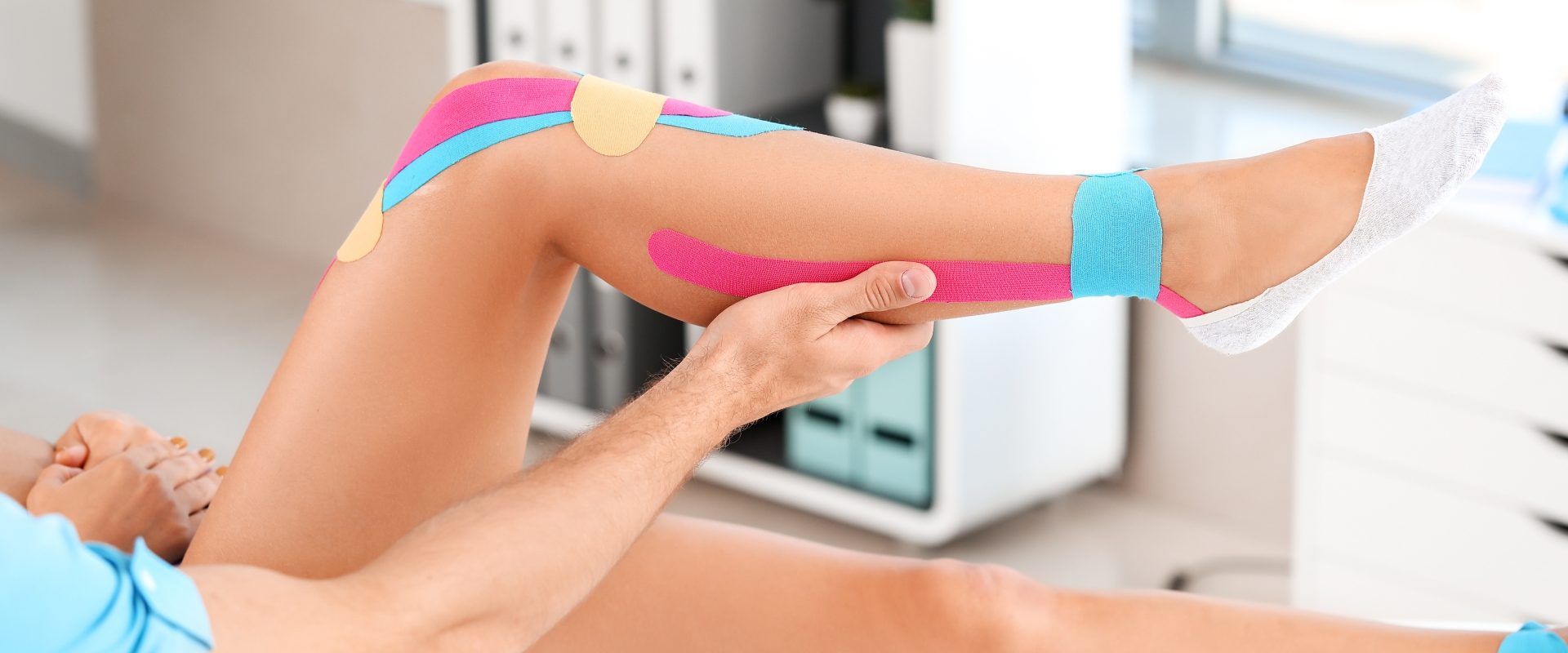 Kinesiotaping - na czym polega? Fizjoterapeuta nakleja pacjentowi kolorowe tejpy.