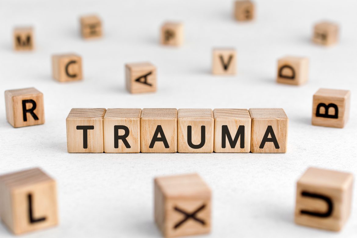 Trauma - czym jest i jak sobie pomóc? Drewniane klocki z czarnymi literkami na białej podłodze ułożone w słowo trauma.