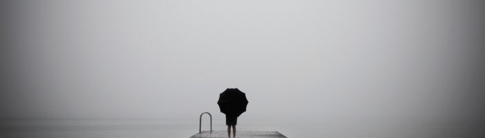 Jak poradzić sobie z traumą? Postać ubrana na czarno, ukryta pod czarnym parasolem stoi na pomoście nad zamglonym i dżdżystym jeziorem i patrzy w dal.