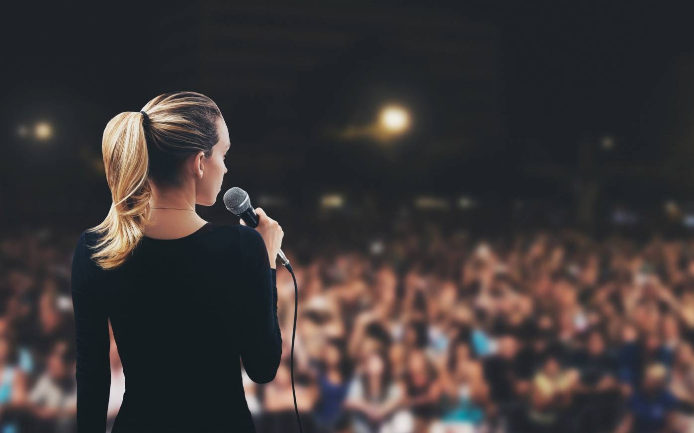 Jak pokonać tremę podczas wystąpienia publicznego? Młoda kobieta z mikrofonem na scenie przemawia przed szeroką publicznością.