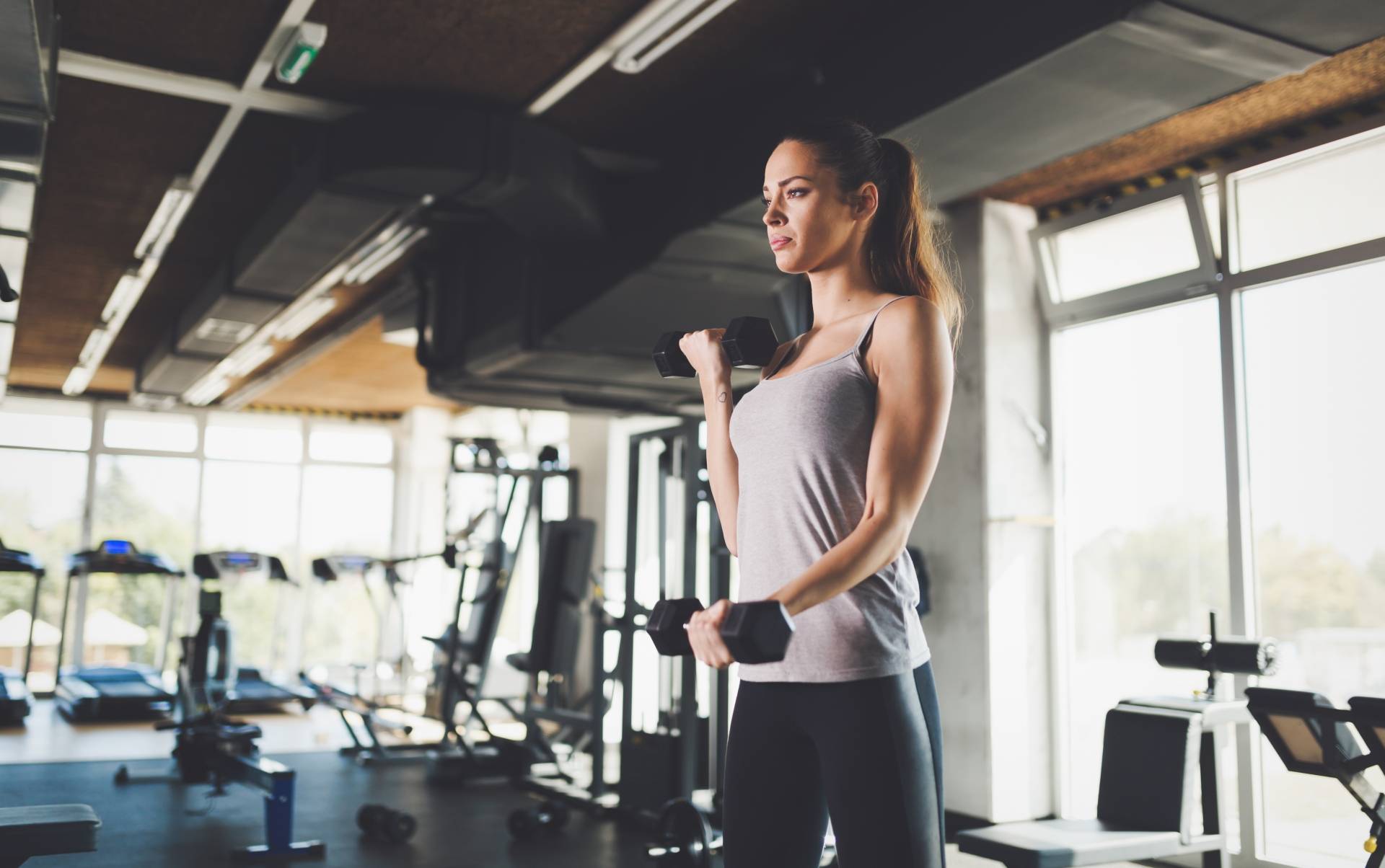 Ćwiczenia na proste plecy - jak wygląda prawidłowa postawa? Kobieta ćwiczy na siłowni z hantlami.
