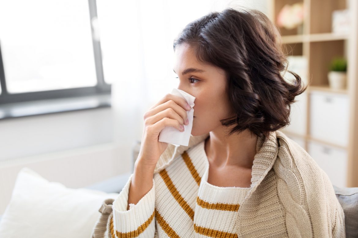 Jak rozpoznać czy to uczulenie czy przeziębienie? Jak odróżnić objawy alergii od przeziębienia? Młoda kobieta siedzi w domu na kanapie i wydmuchuje nos w chusteczkę.