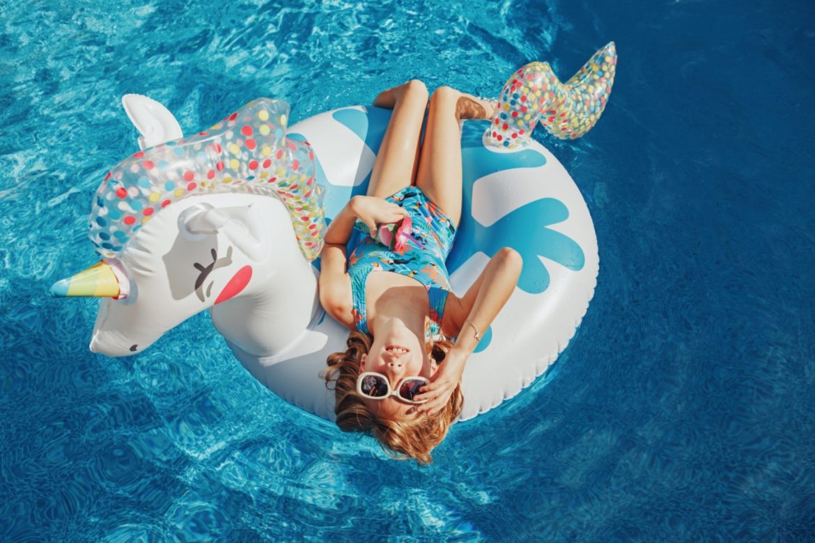 Wakacje z dziećmi - jak je dobrze zaplanować? Dziewczynka leży na materacu dmuchanym w kształcie jednorożca w basenie.