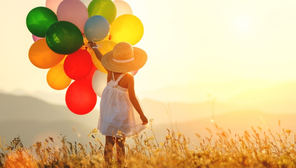 Codzienna praktyka wdzięczności dla dzieci – czyli jak uczyć dziecko szczęśliwego życia? Mała dziewczynka stoi na łące i trzyma w ręku kolorowe balony.