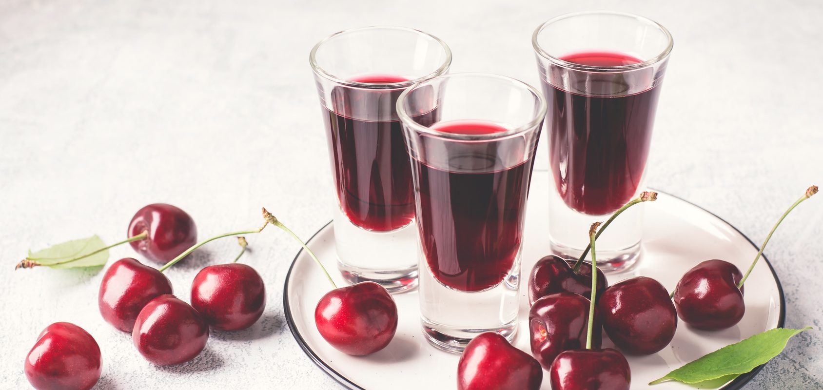 Nalewka wiśniowa - wiśniówka w 3 kieliszkach na białej okrągłej tacy, obok leżą owoce wiśni. Sprawdź przepis na wiśniówkę.