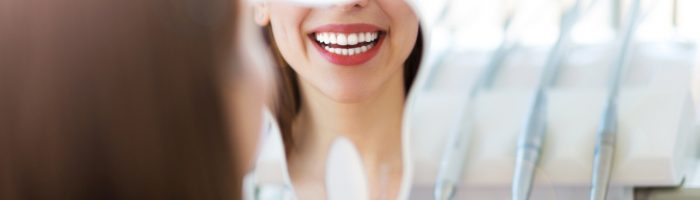 Strach przed dentystą - jak nie bać się dentysty? Kobieta przygląda się swojemu uzębieniu w małym lusterku, siedząc na fotelu w gabinecie dentystycznym.