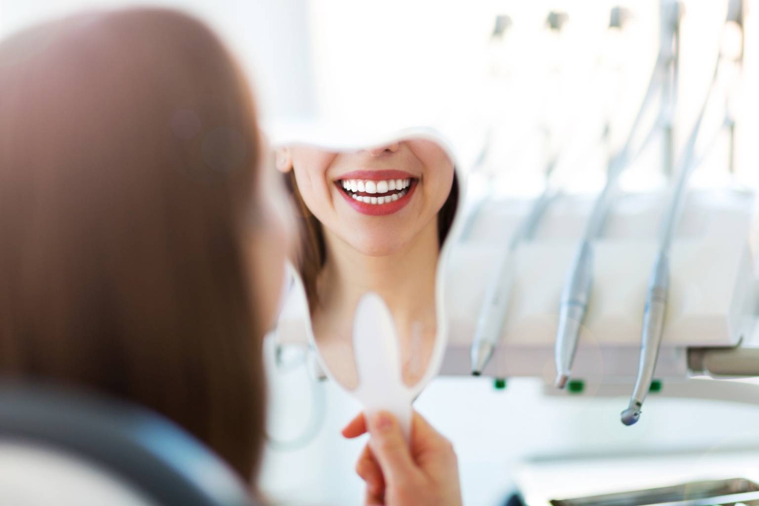 Strach przed dentystą - jak nie bać się dentysty? Kobieta przygląda się swojemu uzębieniu w małym lusterku, siedząc na fotelu w gabinecie dentystycznym.