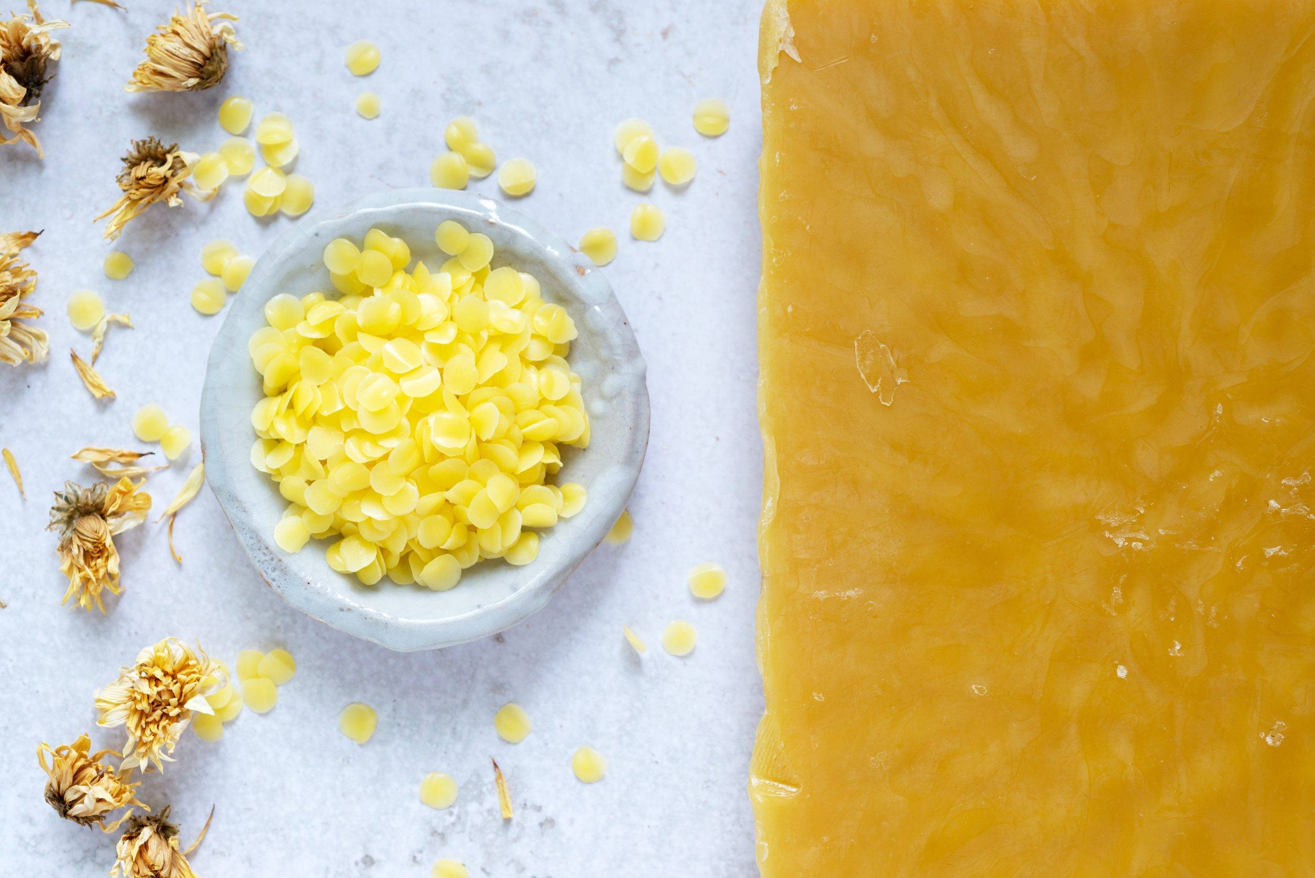 Płatki wosku pszczelego kosmetycznego w miseczce, obok leży plaster wosku. Jak używać wosku pszczelego i jakie ma on właściwości.