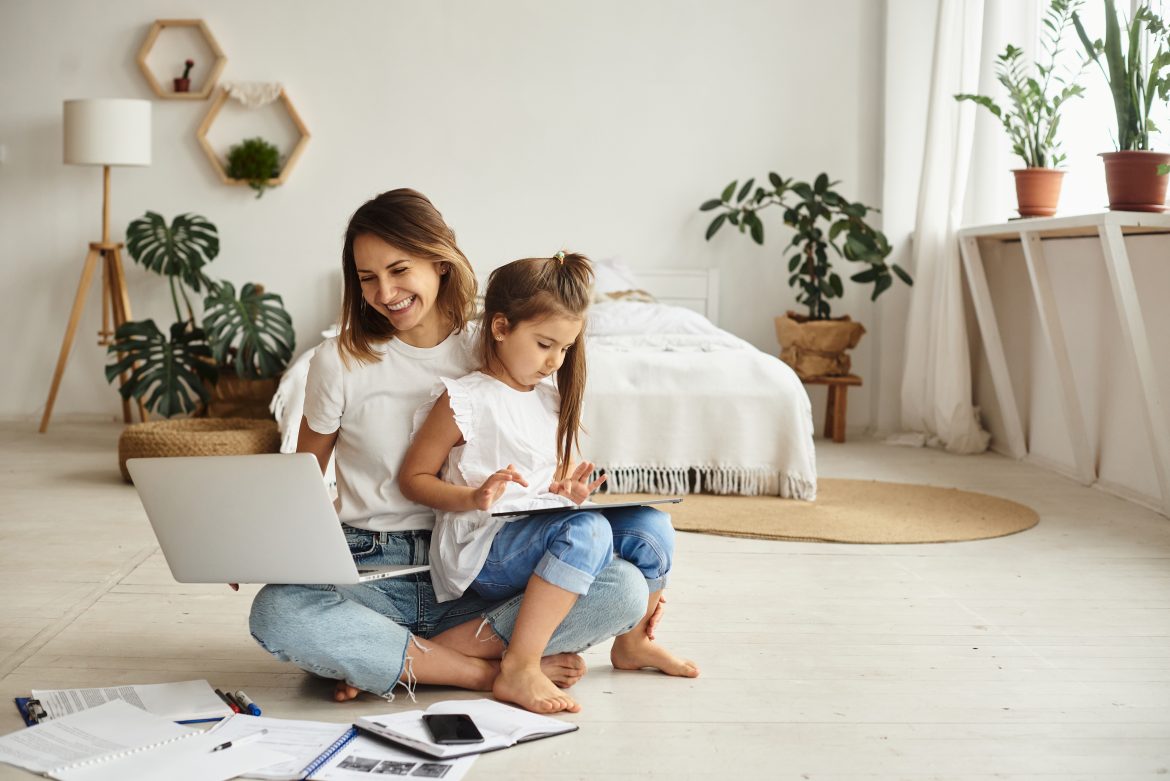 Współczesna mama - czy naprawdę musi być idealna? Mama pracująca w domu - siedzi na podłodze w sypialni, na jednym kolanie trzyma laptopa, na drugim kolanie siedzi jej córka o rysuje.