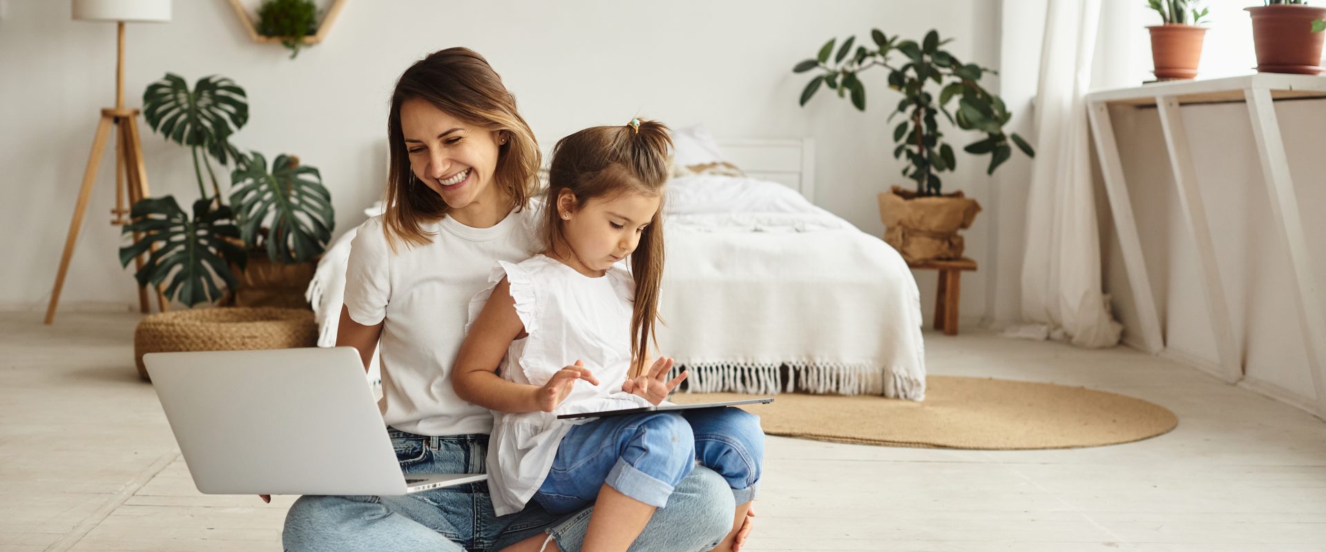 Współczesna mama - czy naprawdę musi być idealna? Mama pracująca w domu - siedzi na podłodze w sypialni, na jednym kolanie trzyma laptopa, na drugim kolanie siedzi jej córka o rysuje.