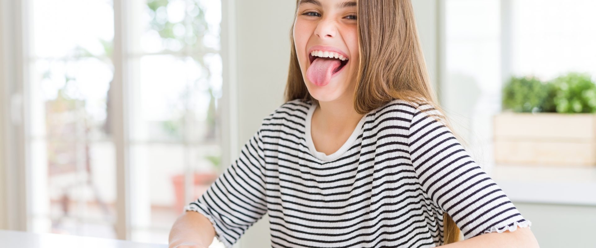 Zmiany na języku - o czym świadczy wygląd języka? Młoda dziewczyna w koszulce w paski siedzi przy blacie kuchennym i pokazuje język.