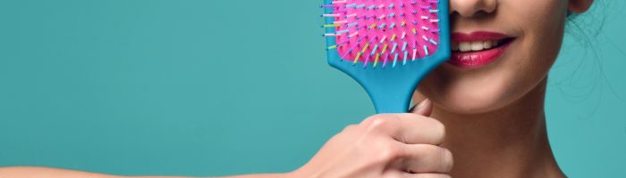 Wypadanie włosów - jakie są przyczyny, jak leczyć łysienie u kobiet naturalnymi metodami? Uśmiechnięta dziewczyna z uwiązanymi do góry włosami przykłada do oka kolorową szczotkę do włosów, stoi na tle niebieskiej ściany.