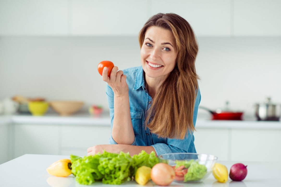 Zakwaszenie organizmu - czym jest i co warto o nim wiedzieć? Kobieta opiera się łokciami o blat kuchenny, trzymając w ręku pomidora.