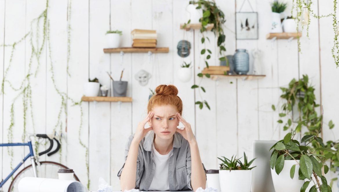 Zarządzanie czasem - jak być efektywną w pracy i w życiu? Rudowłosa dziewczyna siedzi przy biurku i zastanawia się nad projektem, który leży na biurku, za nią wiszą na ścianie półki z roślinami i dekoracjami.