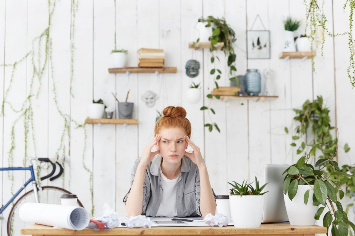 Zarządzanie czasem - jak być efektywną w pracy i w życiu? Rudowłosa dziewczyna siedzi przy biurku i zastanawia się nad projektem, który leży na biurku, za nią wiszą na ścianie półki z roślinami i dekoracjami.