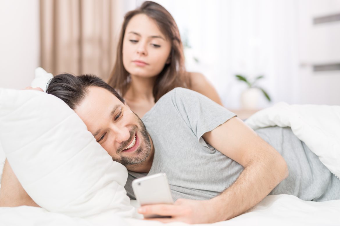 Jak radzić sobie z zazdrością? Kobieta zagląda partnerowi przez ramię, gdy ten patrzy w telefon, leżąc w łóżku.
