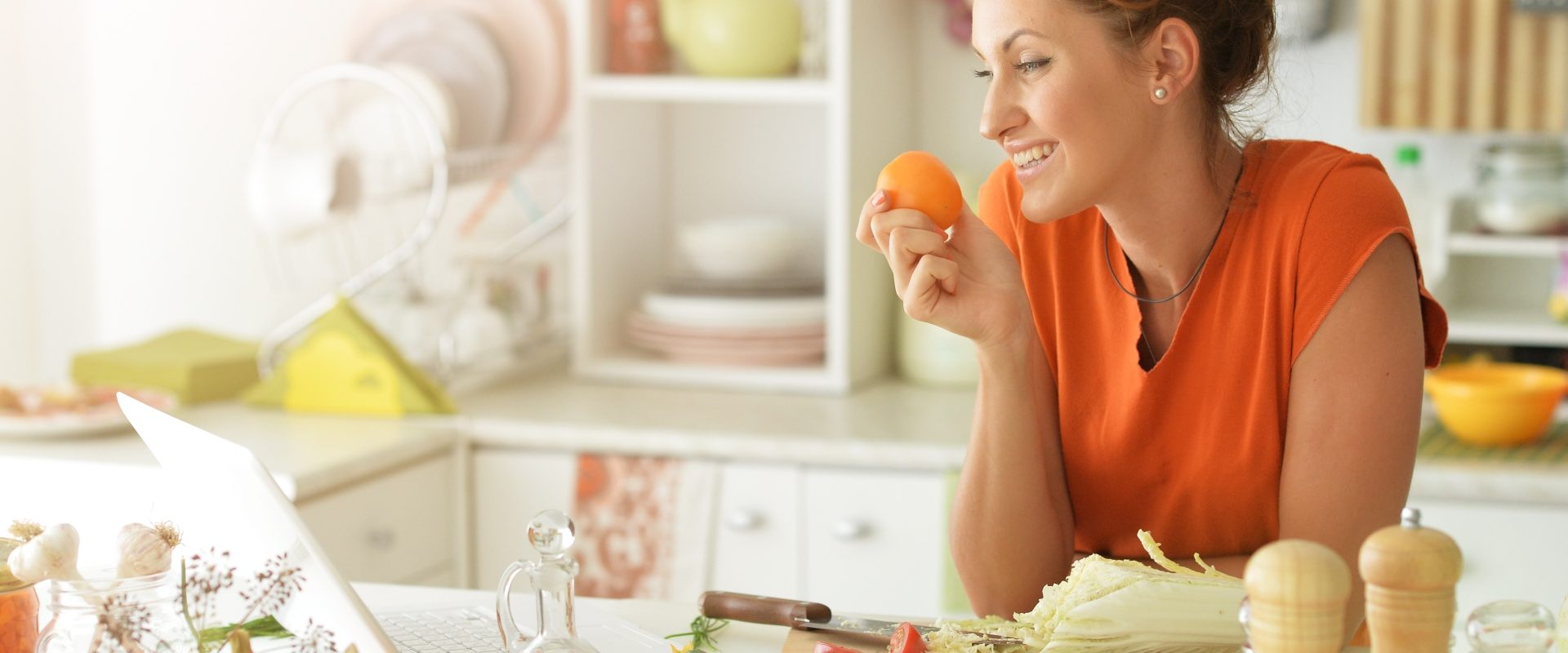 Naturalne sposoby na oczyszczenie wątroby. Kobieta w pomarańczowej bluzce opiera się łokciami o blat w kuchni i trzyma w dłoni mandarynkę. Wokół rozłożone warzywa i owoce.