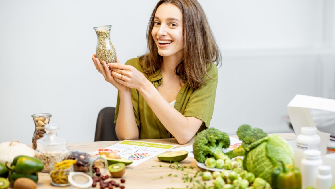 Konopie siewne na talerzu - jak je wykorzystać w kuchni? Uśmiechnięta kobieta w zielonej bluzce siedzi przy stole zastawionym warzywami i owocami i trzyma w ręku szklaną karafkę z nasionami konopi.
