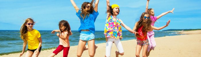 Zatrucie pokarmowe u dzieci - jak je leczyć? Szczęśliwe dzieci w kolorowych ubraniach pozują do zdjęcia na plaży podczas wakacji, skacząc do góry.