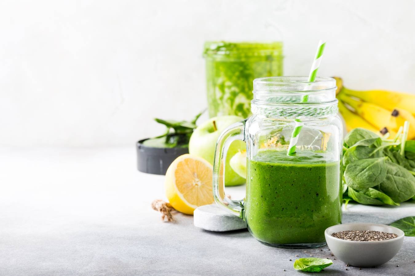 Zielony koktajl na odporność - źródło chlorofilu i witamin. Słoik ze smoothie z zielonych warzyw i owoców stoi na blacie, wokół leżą składniki: szpinak, nasiona, cytryna, zielone jabłko, banany.