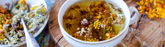 Leki roślinne na uspokojenie i dobry sen. Filiżanka ziołowej herbaty na drewnianej desce i błękitnym rustykalnym stole, obok leży spodeczek, a wokół rozrzucone ususzone zioła i kwiaty.