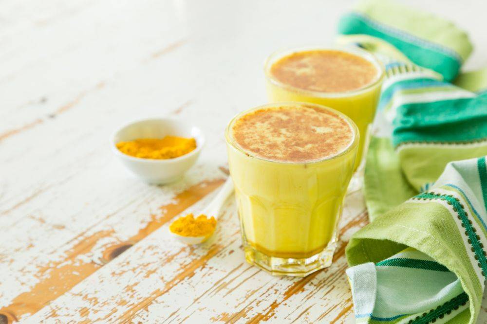 Złote mleko na przeziębienie. Napój z kurkumy rozlany do dwóch kieliszków, które stoją na białym rustykalnym blacie, obok leży miseczka z kurkumą i zielona ściereczka kuchenna.