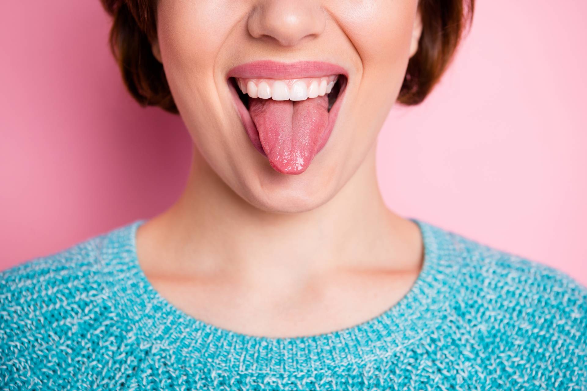 O czym świadczą zmiany na języku? Młoda dziewczyna w niebieskim swetrze wystawia język. Stoi na tle różowej ściany. 