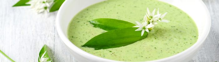 Przepisy z czosnkiem niedźwiedzim Agnieszki Żelazko. Zupa krem z białych warzyw w białej misce na jasnym blacie, wokół leżą liście i kwiaty dzikiego czosnku.